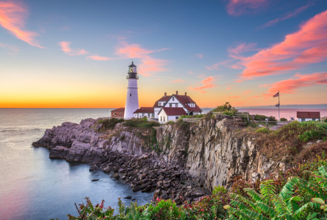 small home sits on a cliff near the seaside with a lighthouse nearby at sunset