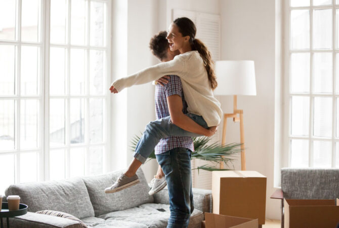Woman lifted off ground with legs wrapped around man surrounded by moving boxes in their apartment