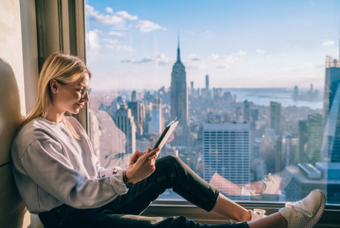 Girl reading next to window overlooking skyscrapers.