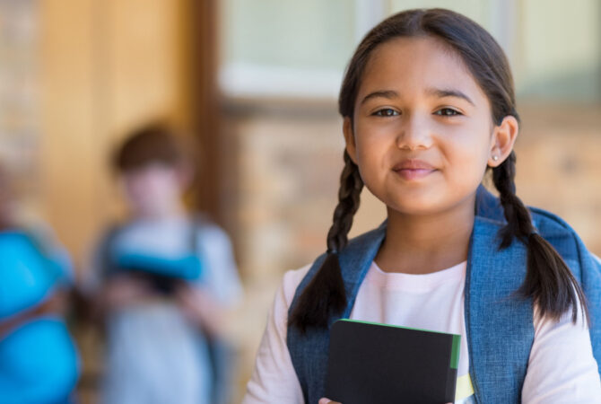 Young girl standing in school holding a notebook and carrying backpack.