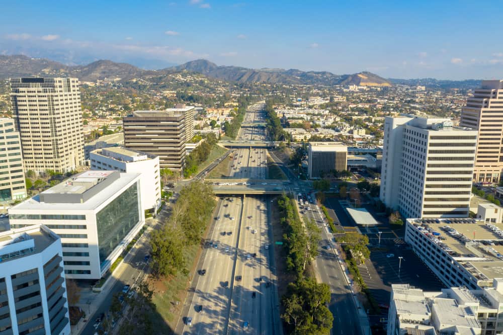 Glendale, CA drone shot of highway lined by trees and tall buildings on clear day with mountains in background
