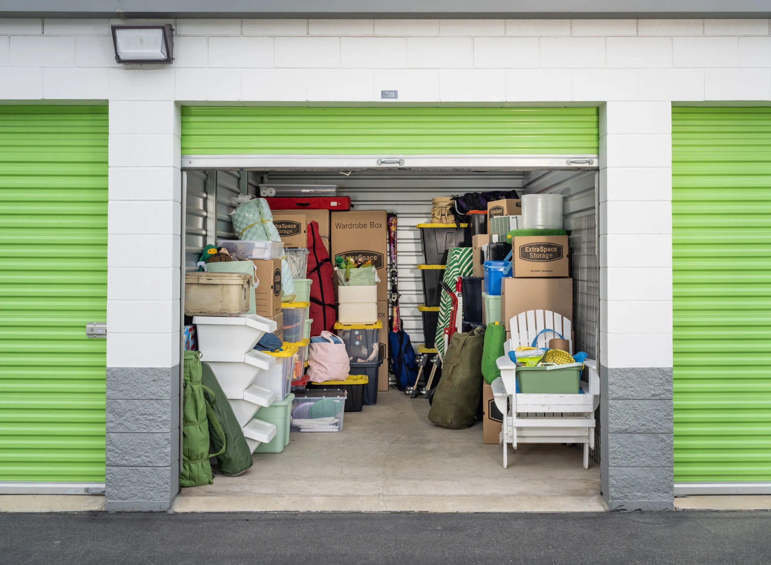 Extra Space Storage unit with green garage door open and filled with storage bins, cardboard boxes, and furniture